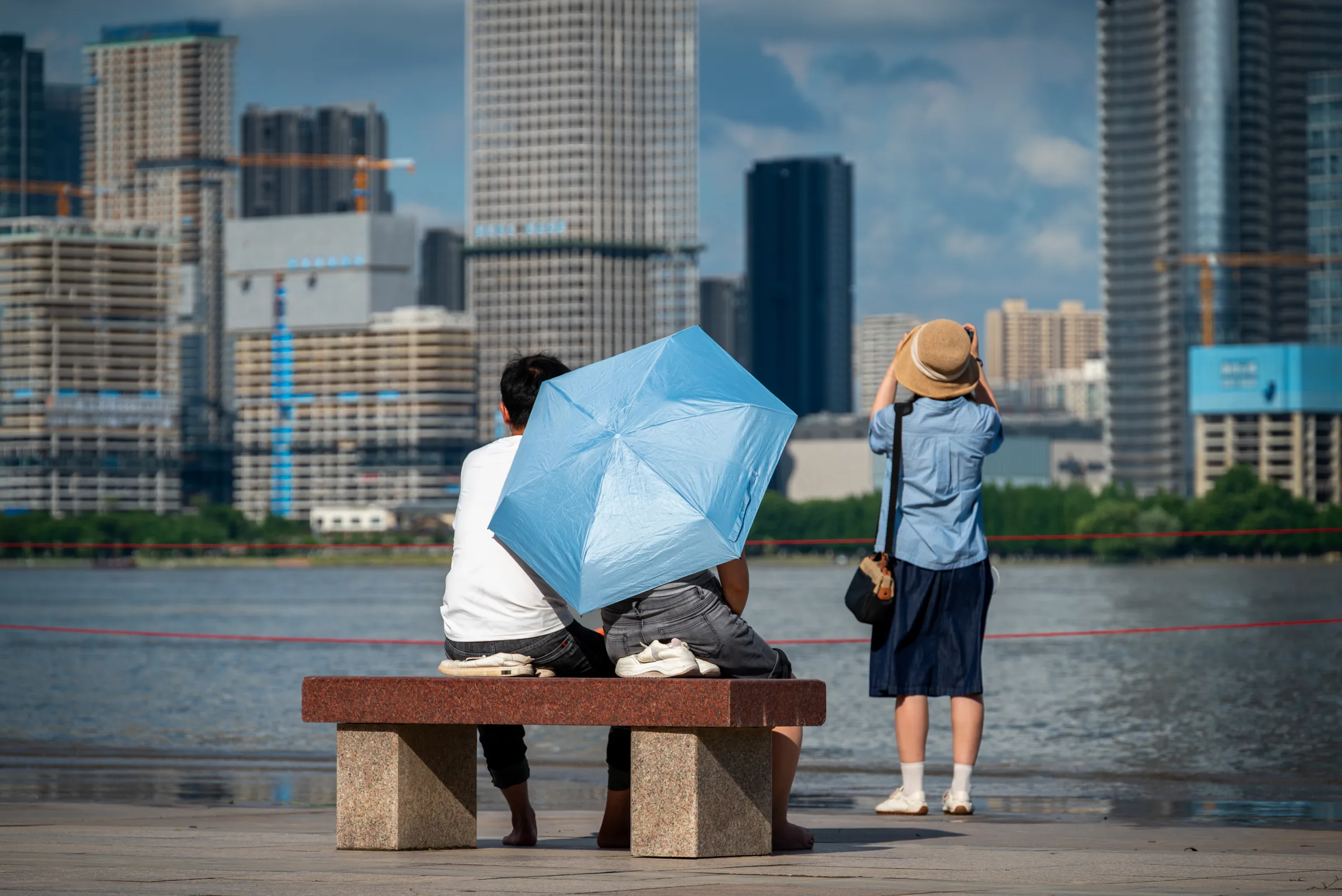 People under a blue umbrella on a riverside bench, with towers behind