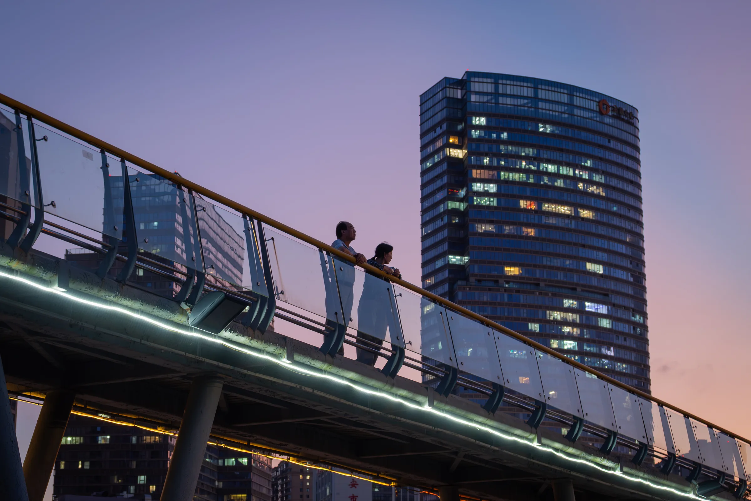 Two people on a curved footbridge at dusk beside a lit tower